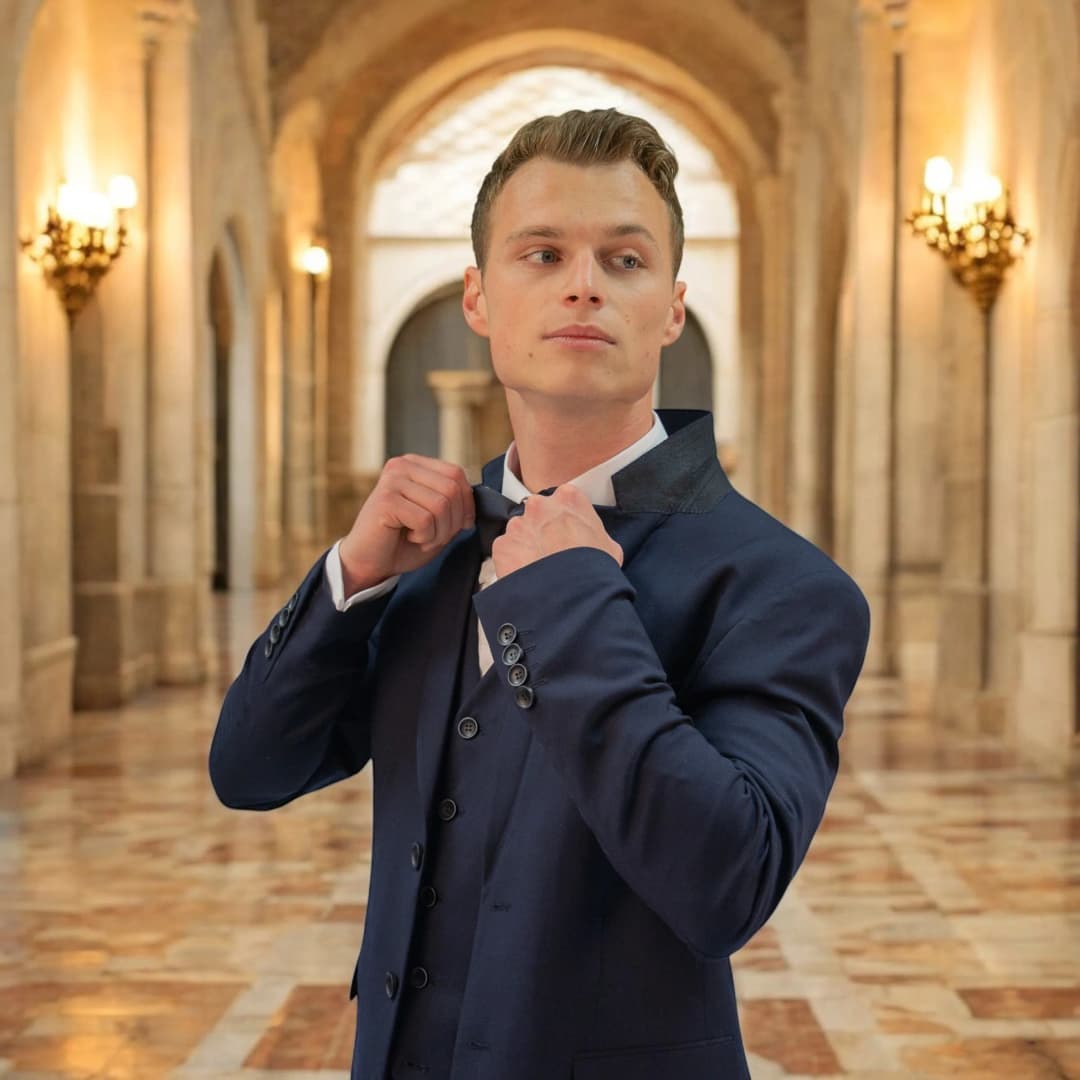 Homme en costume trois pièces bleu marine qui ajuste son nœud papillon dans une galerie à arcades éclairée par des appliques