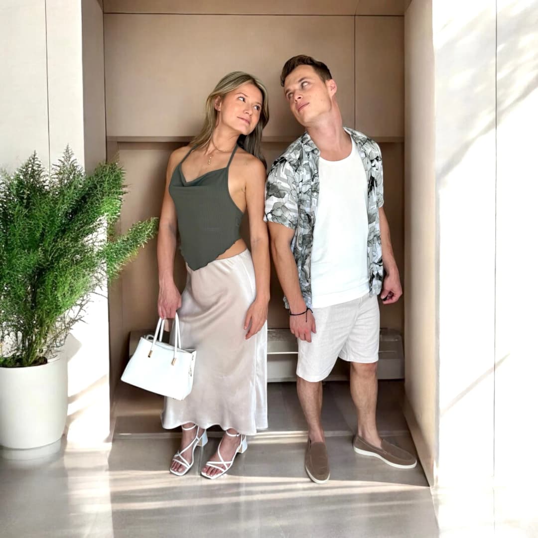 Couple debout dans une niche claire, elle en top dos nu kaki et jupe satinée beige avec un sac blanc, lui en chemise imprimée ouverte sur un t-shirt blanc et bermuda clair
