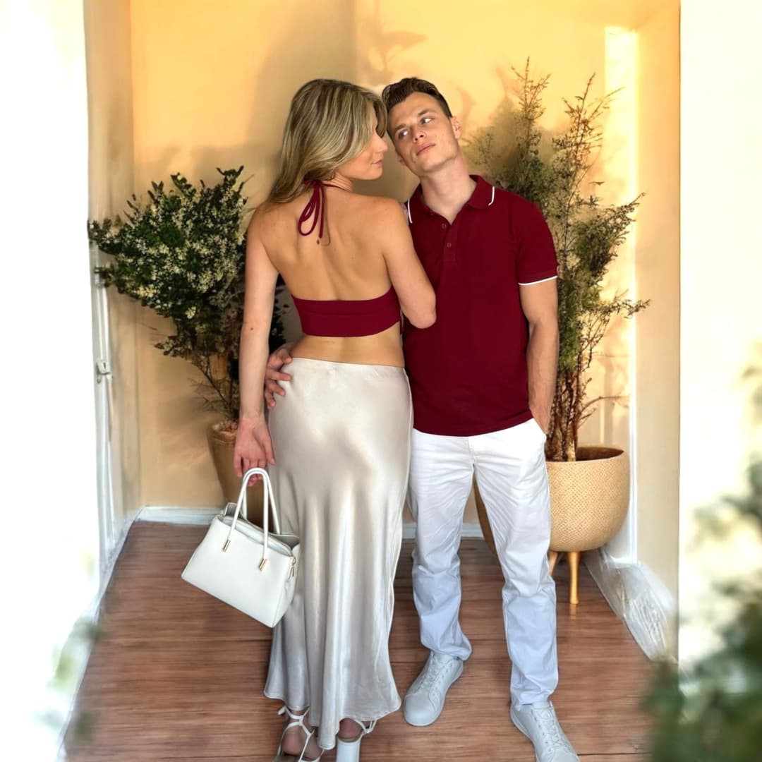 Couple debout entre deux plantes, elle de dos en top dos nu bordeaux et jupe longue satinée beige, lui en polo bordeaux et pantalon blanc
