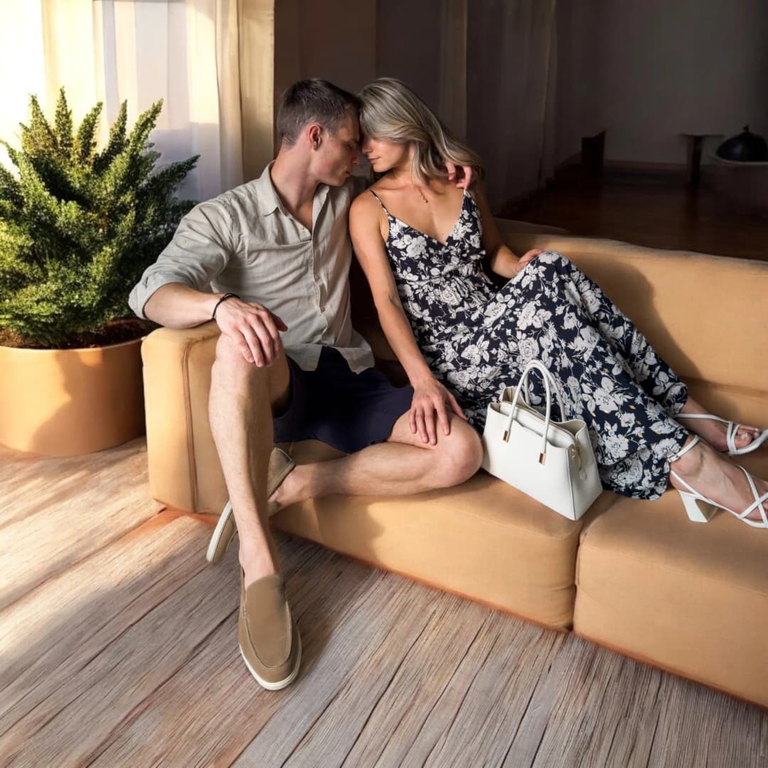 Couple assis sur un canapé ocre, elle en robe longue à fleurs blanches sur fond bleu marine, lui en chemise de lin beige et bermuda marine