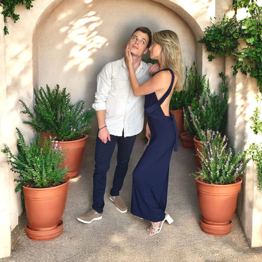 Couple dans une allée bordée de pots en terre cuite, elle en robe longue bleu marine dos nu, lui en chemise à manches longues et pantalon bleu marine