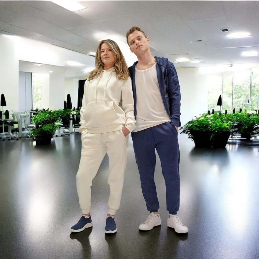 Couple debout dans une salle de sport, elle en jogging à capuche blanc et baskets bleu marine, lui en jogging bleu marine et baskets blanches