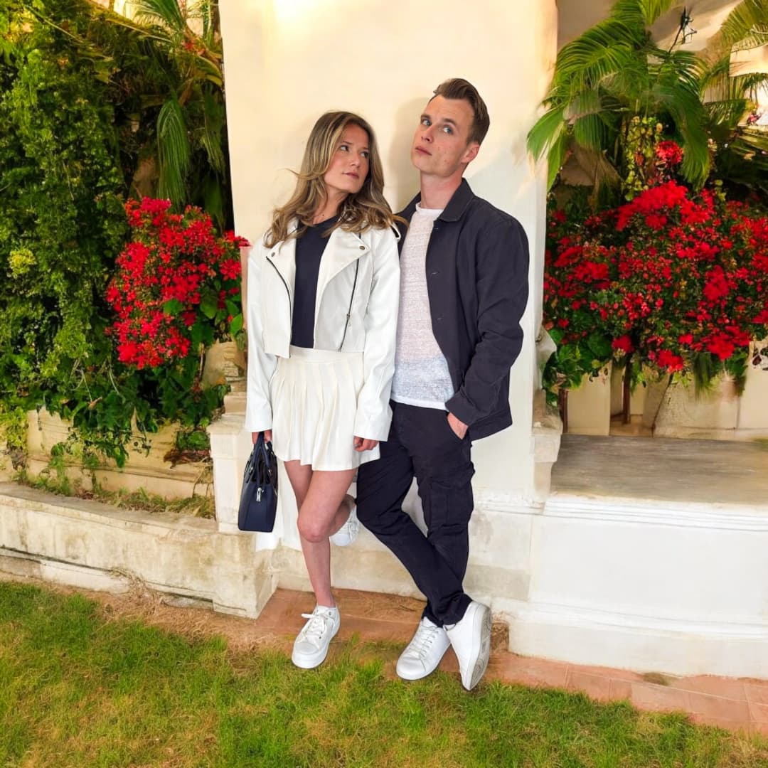 Couple devant un mur blanc et des fleurs rouges, elle en perfecto blanc, haut marine et jupe plissée blanche, lui en veste foncée, t-shirt clair et pantalon foncé