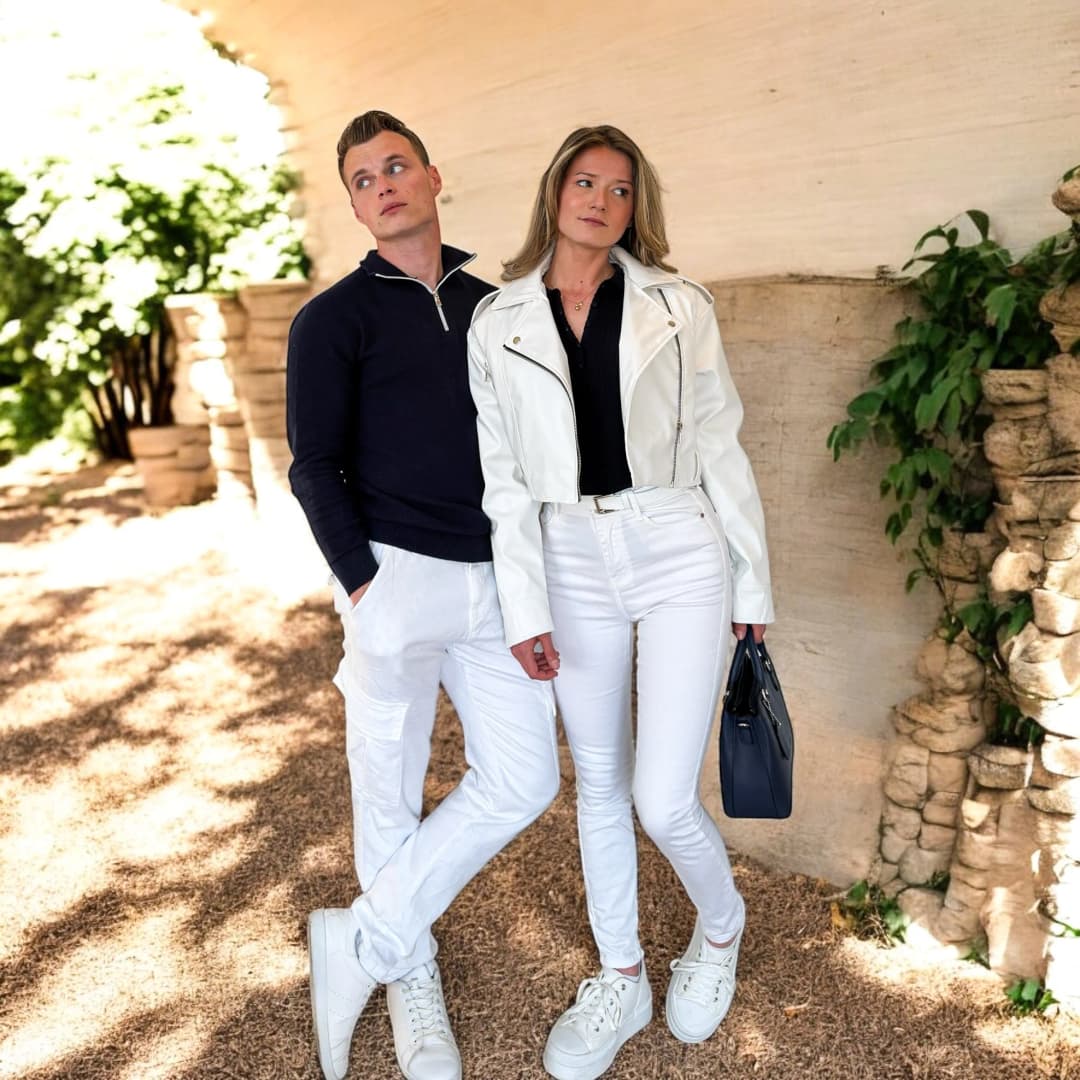 Couple adossé à un mur de pierre, lui en pull zippé bleu et pantalon blanc, elle en perfecto blanc et jean blanc, tous deux en baskets blanches