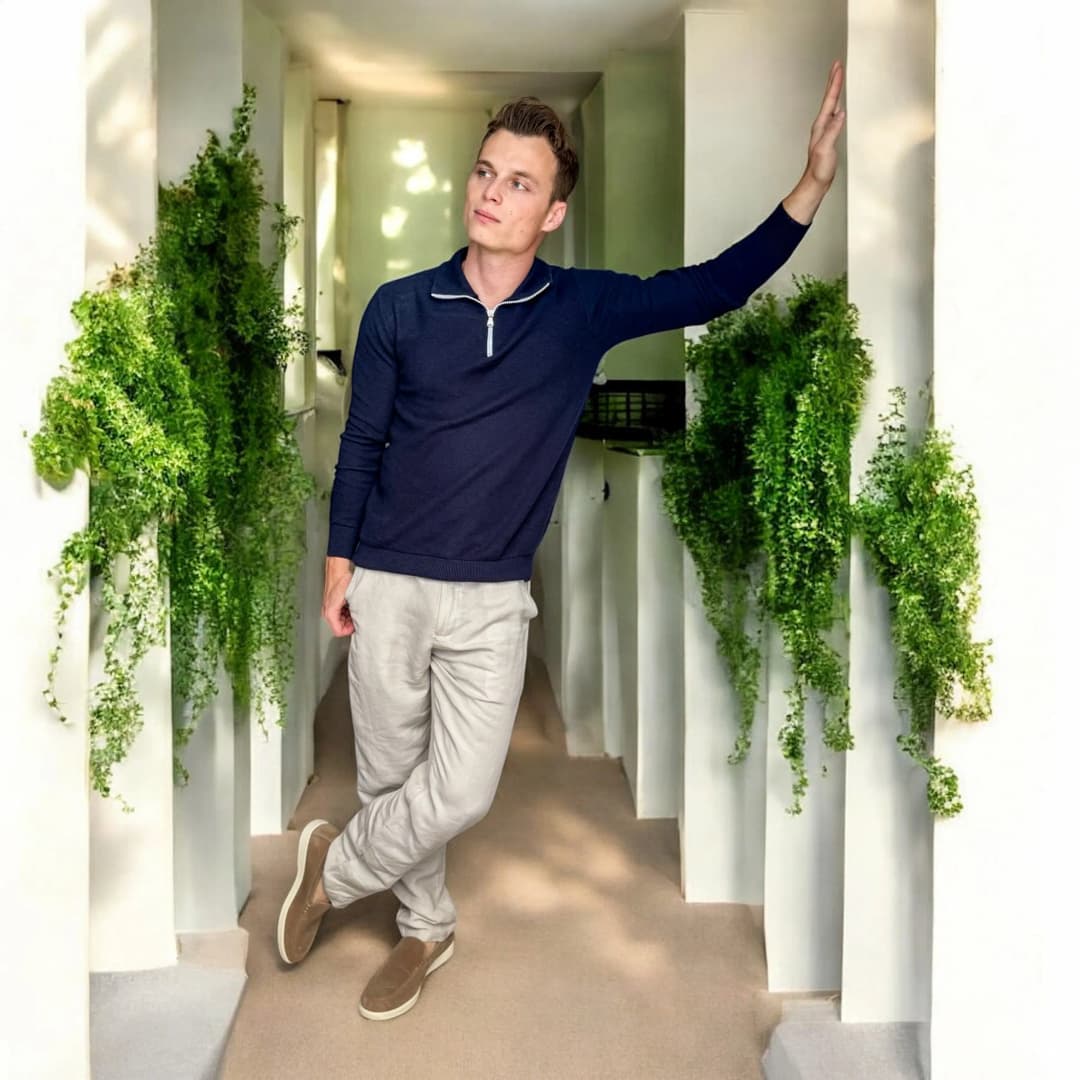 Homme adossé à un mur blanc entre des plantes grimpantes, en pull bleu marine zippé, pantalon en lin clair et mocassins beiges