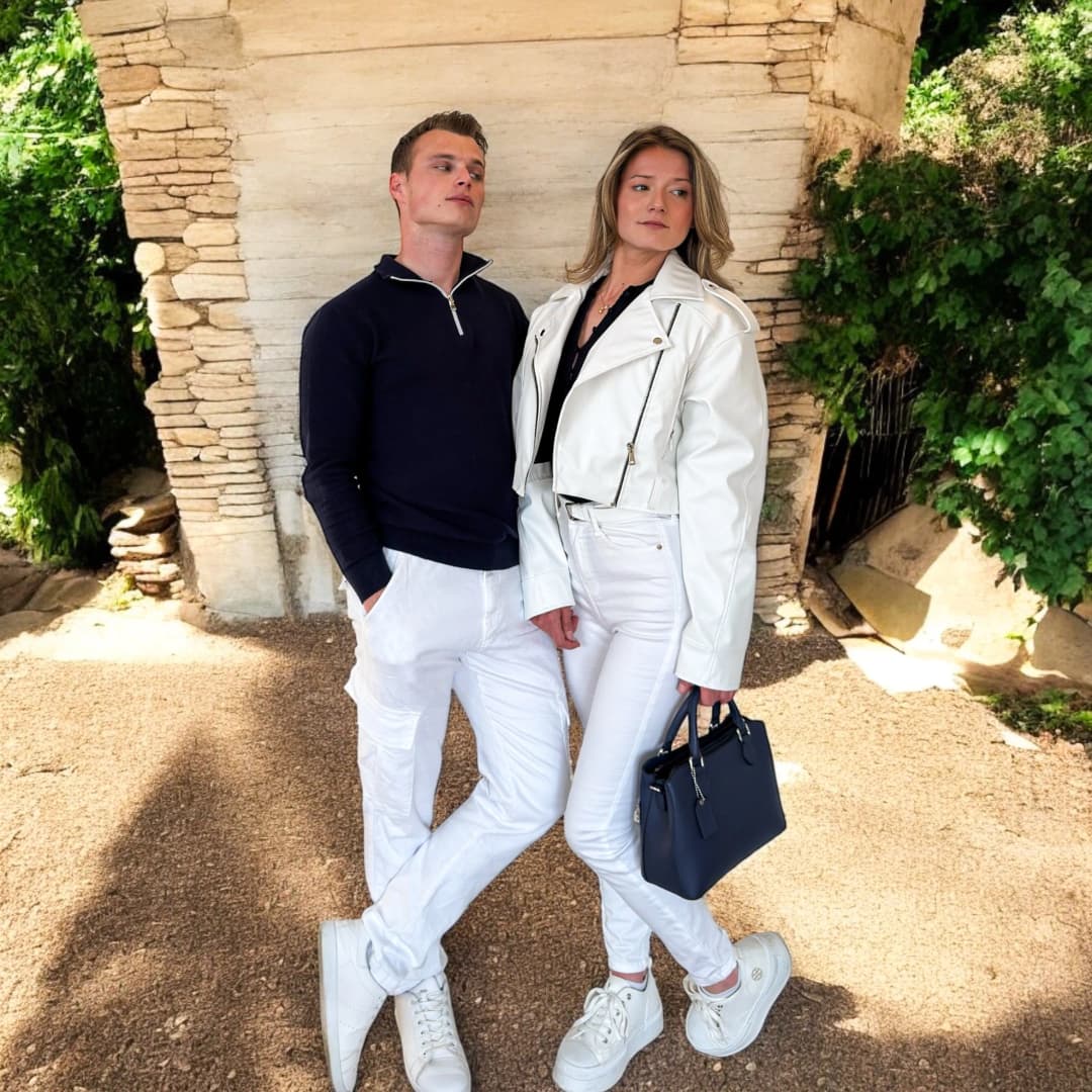 Couple adossé à un mur de pierre, lui en pull zippé bleu marine et pantalon blanc, elle en blouson court blanc, jean blanc et sac bleu marine